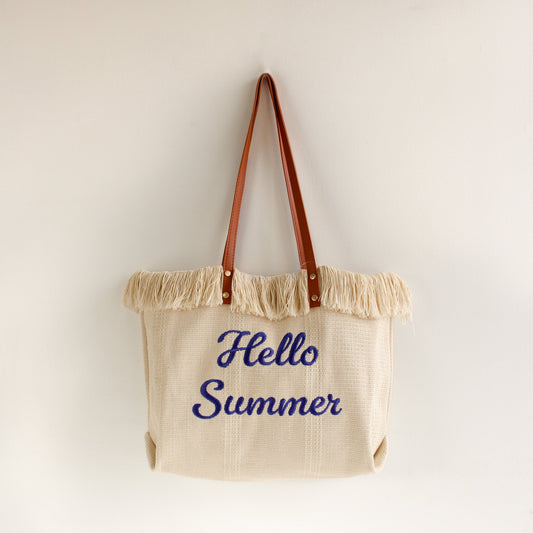 White tote bag with brown leather handles and fringe trim, featuring blue embroidered “Hello Summer” text — a stylish and playful accessory perfect for summer!